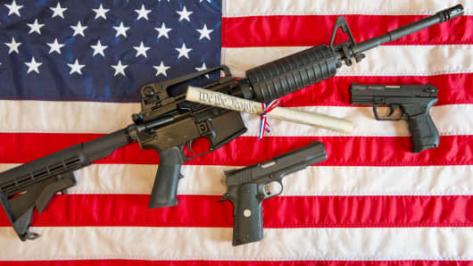 A Colt AR-15 semi-automatic rifle, a Colt .45 semi-auto handgun and a Walther PK380 semi-auto handgun and a copy of the US Constitution on top of an American flag.