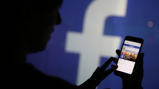 A woman checks the Facebook Inc. site on her smartphone whilst standing against an illuminated wall bearing the Facebook Inc. logo