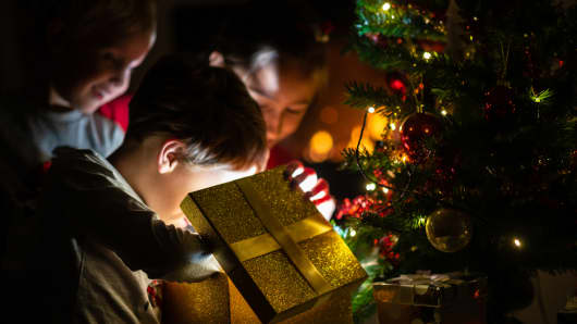 Three kids, two toddler boys and a girl, opening a golden gift box with light coming out of it under a Christmas tree with holiday lights.